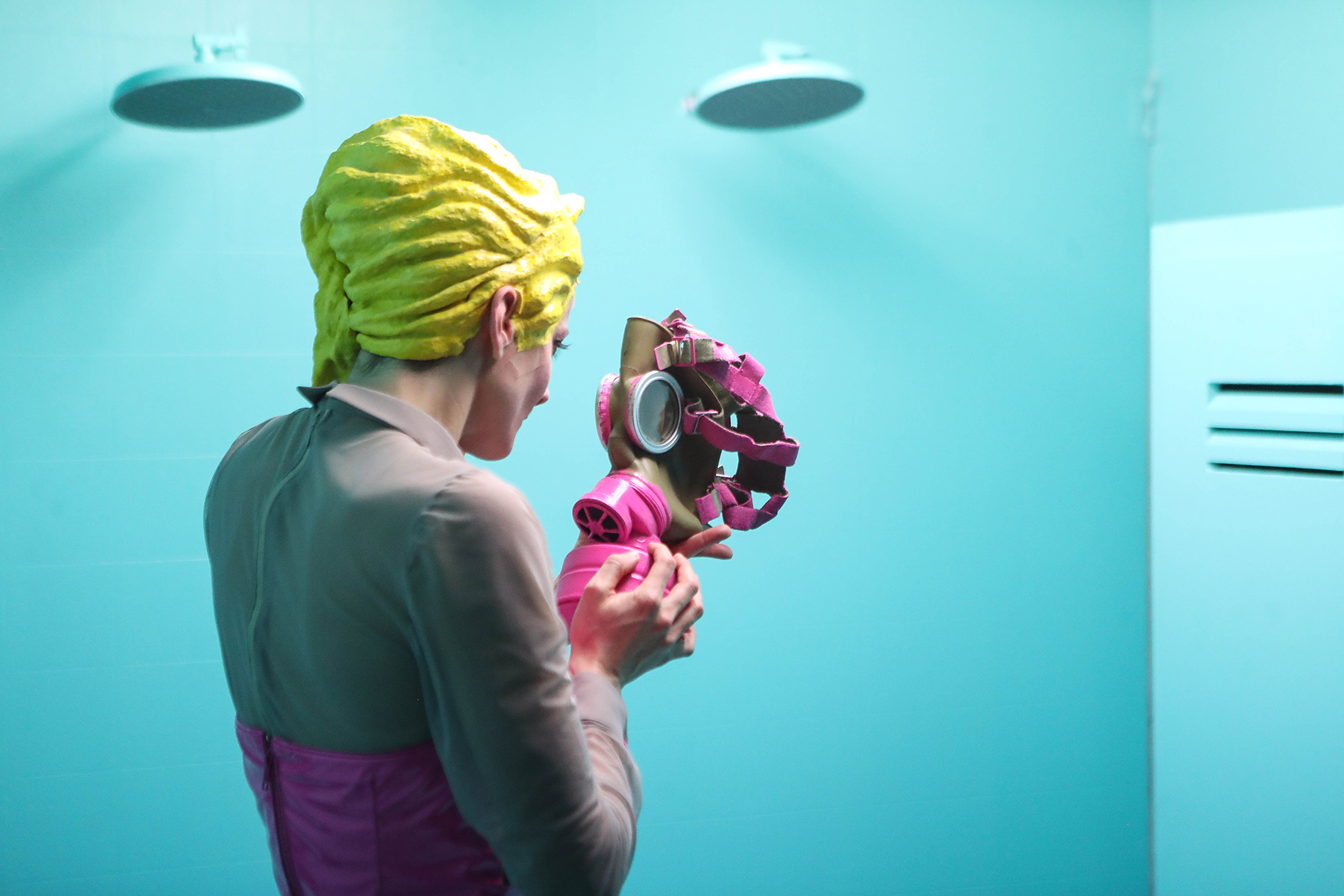 Eine Frau mit gelber Perücke und pinker Kleidung hält eine pinke Gasmaske in der Hand