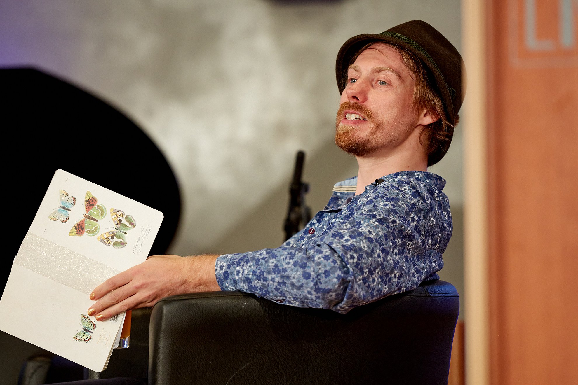 Junger Mann mit Bart und Tirolerhut hat ein Buch über Schmetterlinge in der Hand und spricht